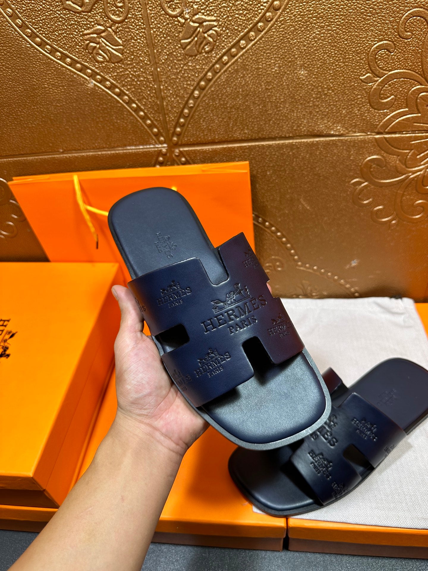 Hand holding Hermes dark blue slide sandals with embossed logo strap and footbed.