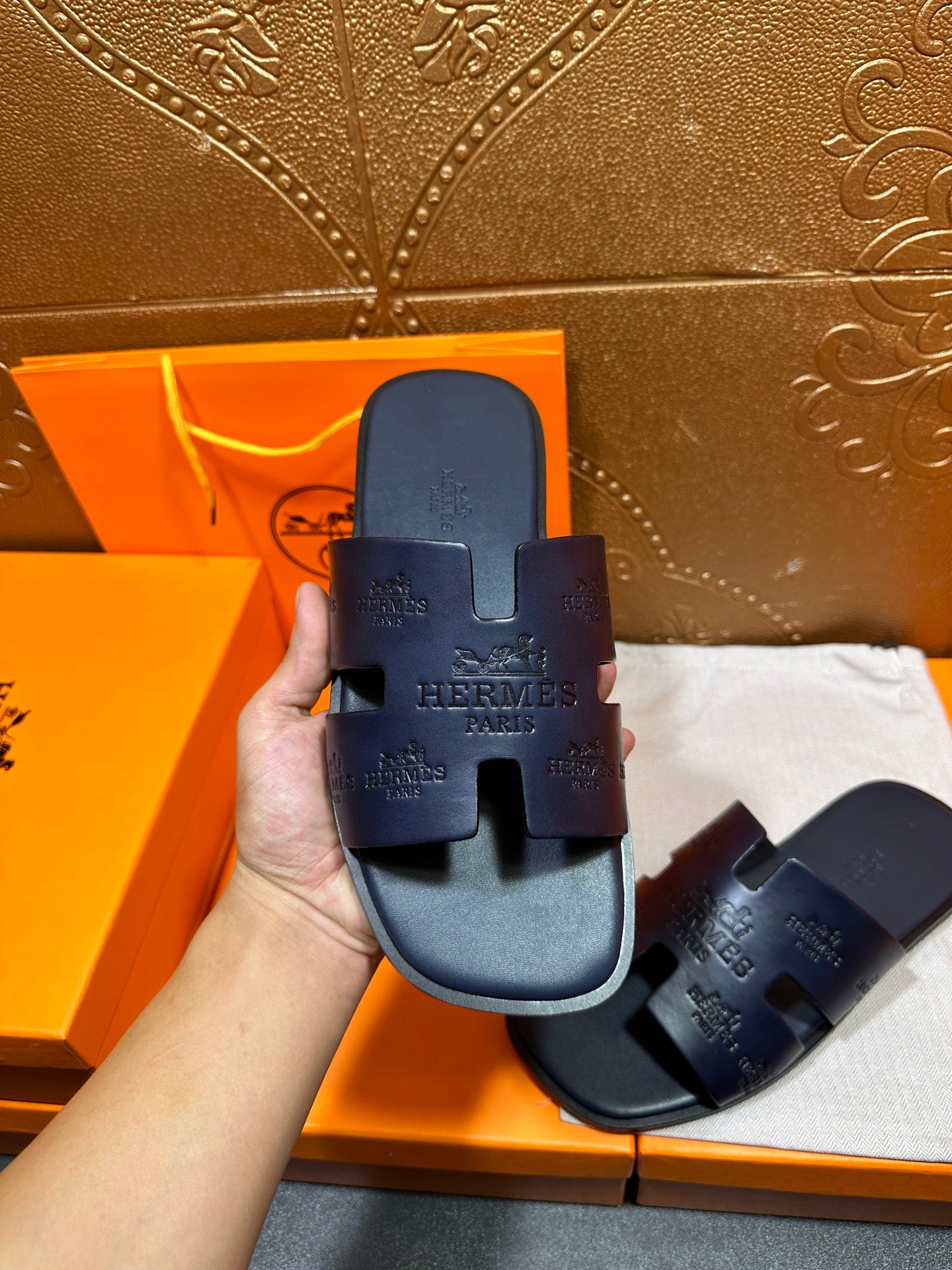 Hand holding Hermes dark blue slide sandals, embossed H logo on strap and footbed.