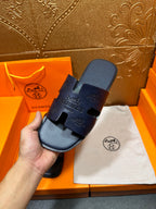 Hand holding Hermes dark blue slide sandals with embossed branding across the strap.