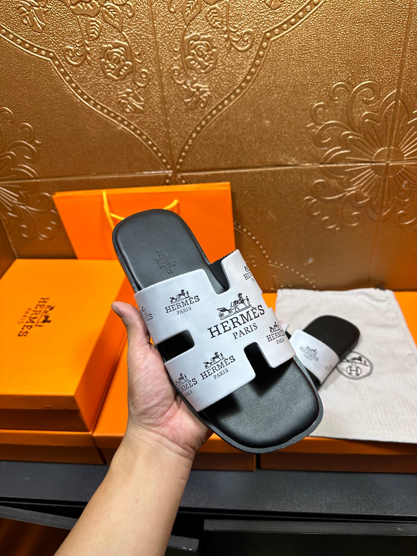 Hand holding Hermes slide sandal with dark gray footbed and white logo strap