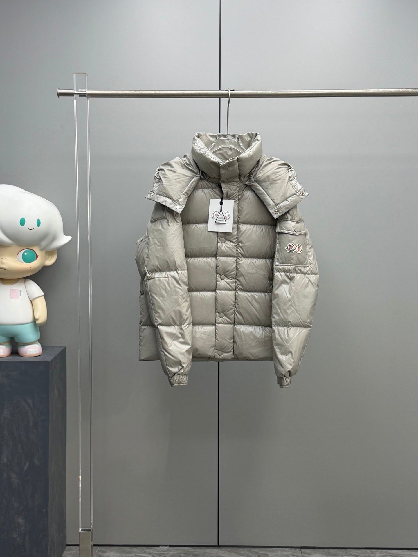 Light gray Moncler puffer jacket with hood and front zipper, hanging on display