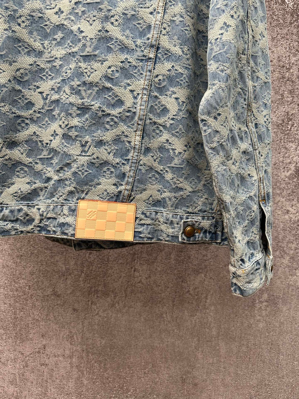 Blue denim jacket with monogram pattern, tan check patch at waist, and gold button