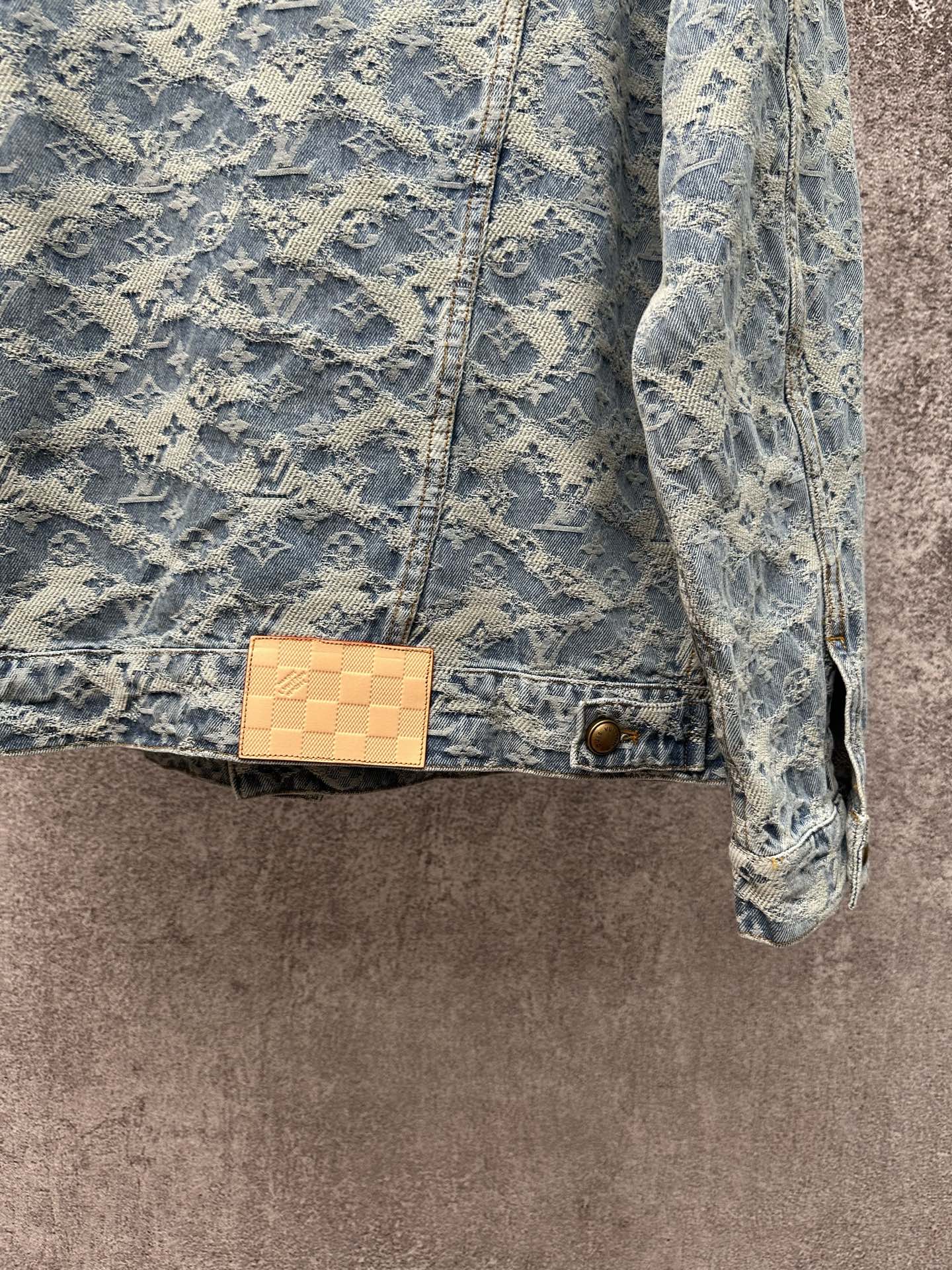 Blue denim jacket with monogram pattern, tan check patch at waist, and gold button