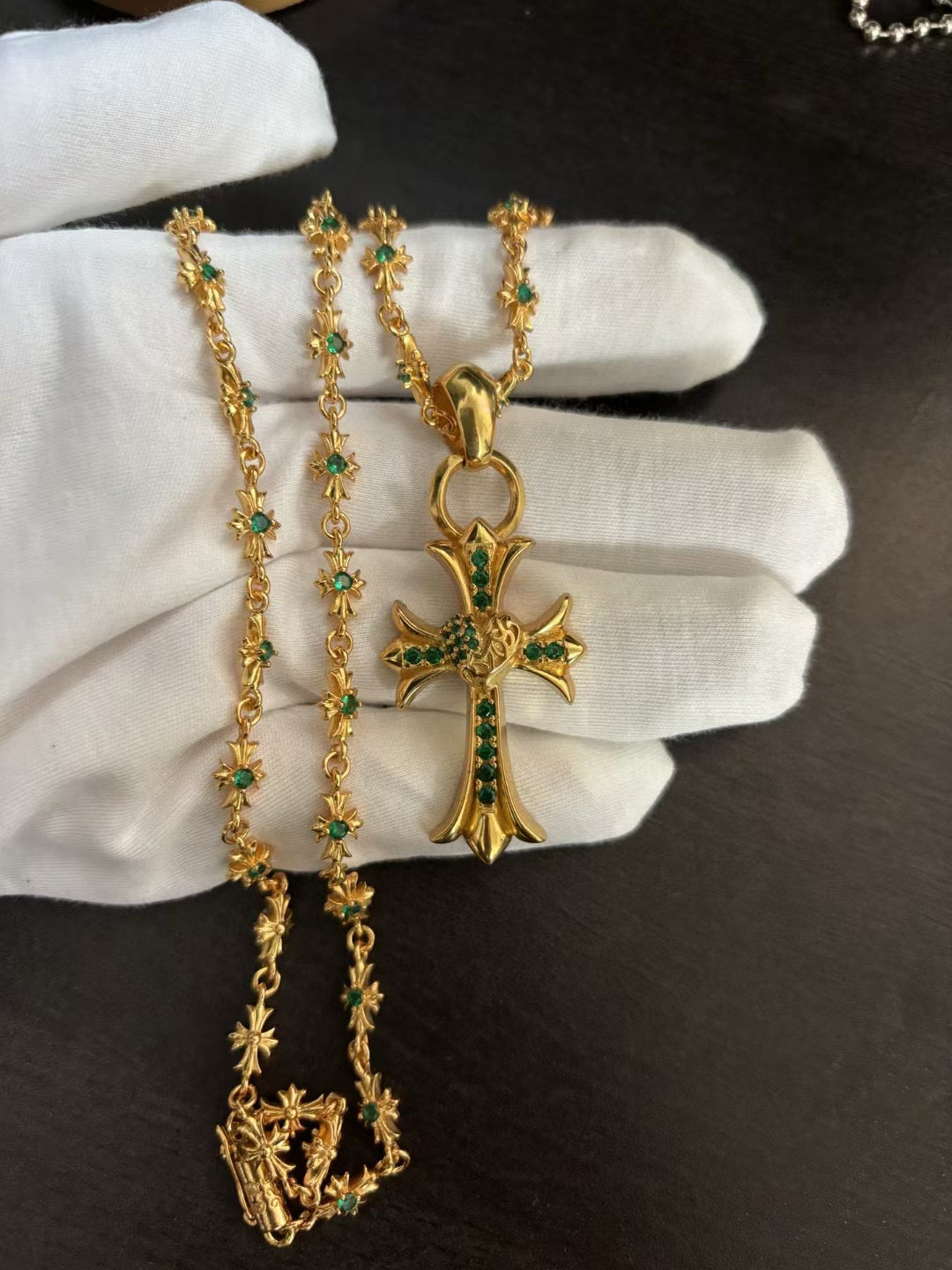 Gold-tone cross pendant necklace with green stones on chain, shown on white gloves