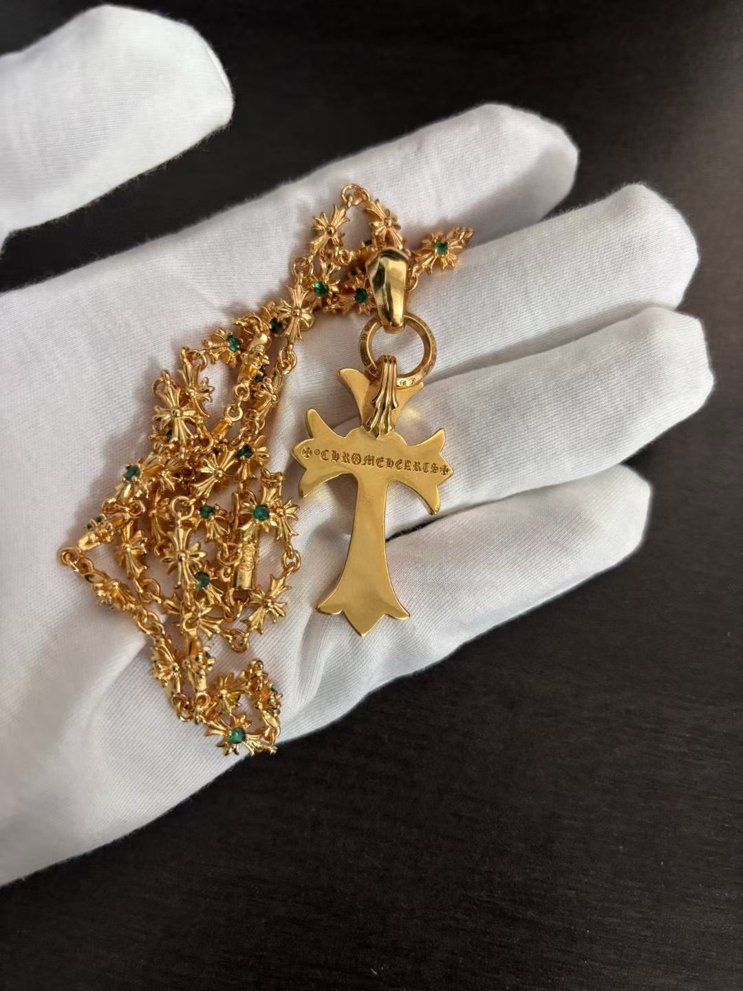 Gold-tone cross pendant with green stones and ornate chain, held on white gloves