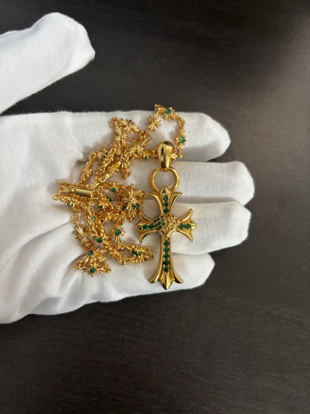 Gold-tone cross pendant necklace with green stone detail, ornate chain on white gloves