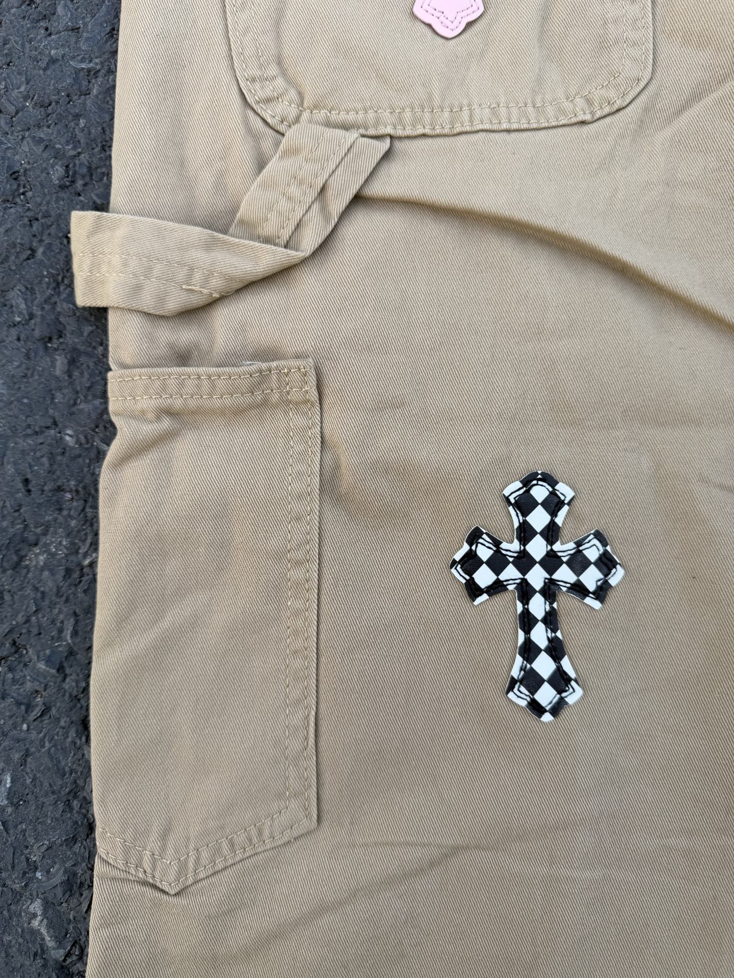 Beige pants with front pocket and black-checkered cross applique near hip