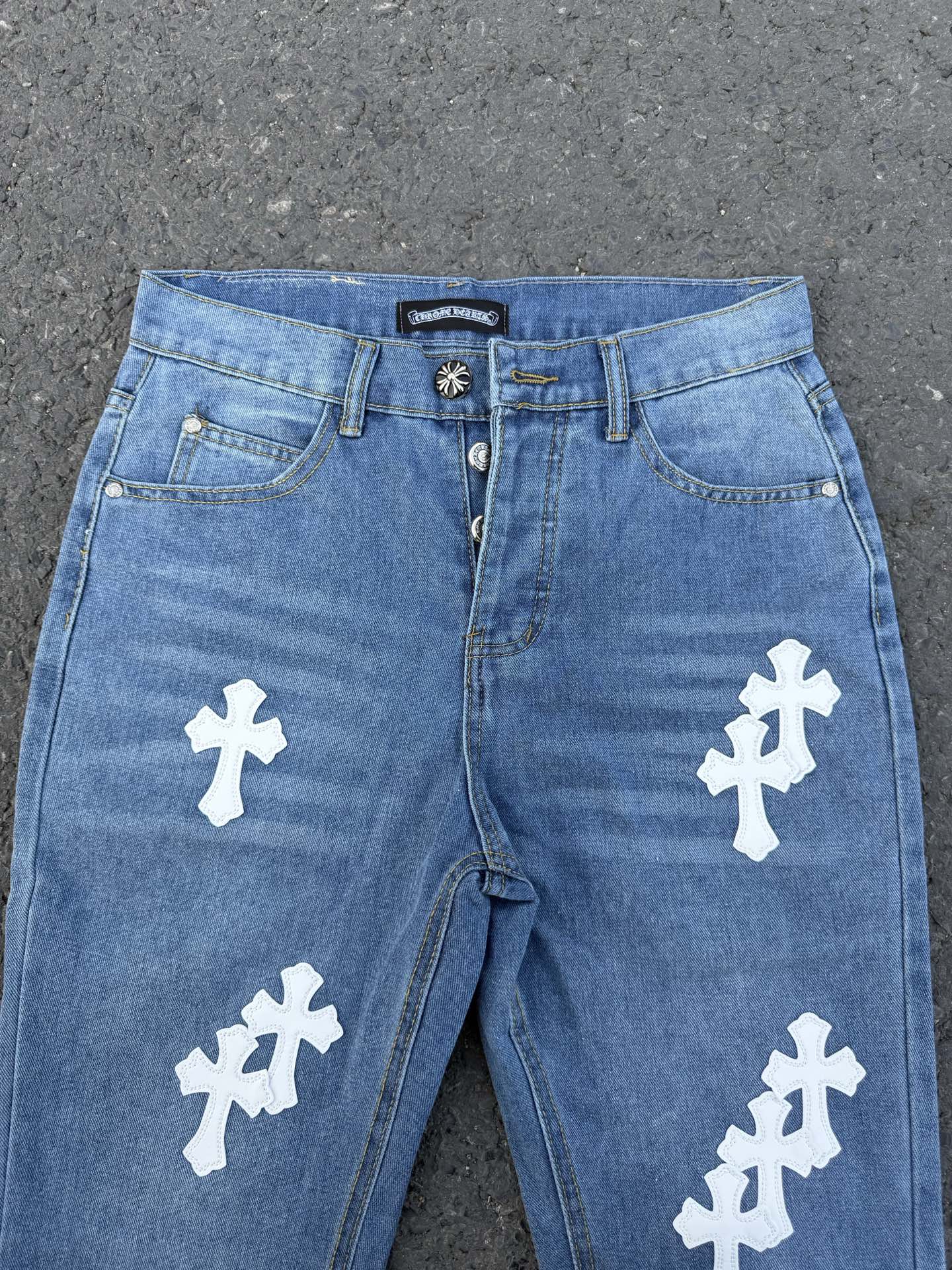 Light blue denim jeans with white cross patches, front waistband view.
