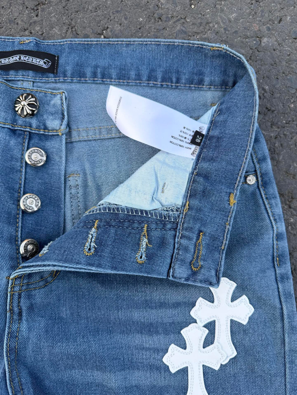 Light blue denim jeans with button fly, distress stitching, white cross patch.