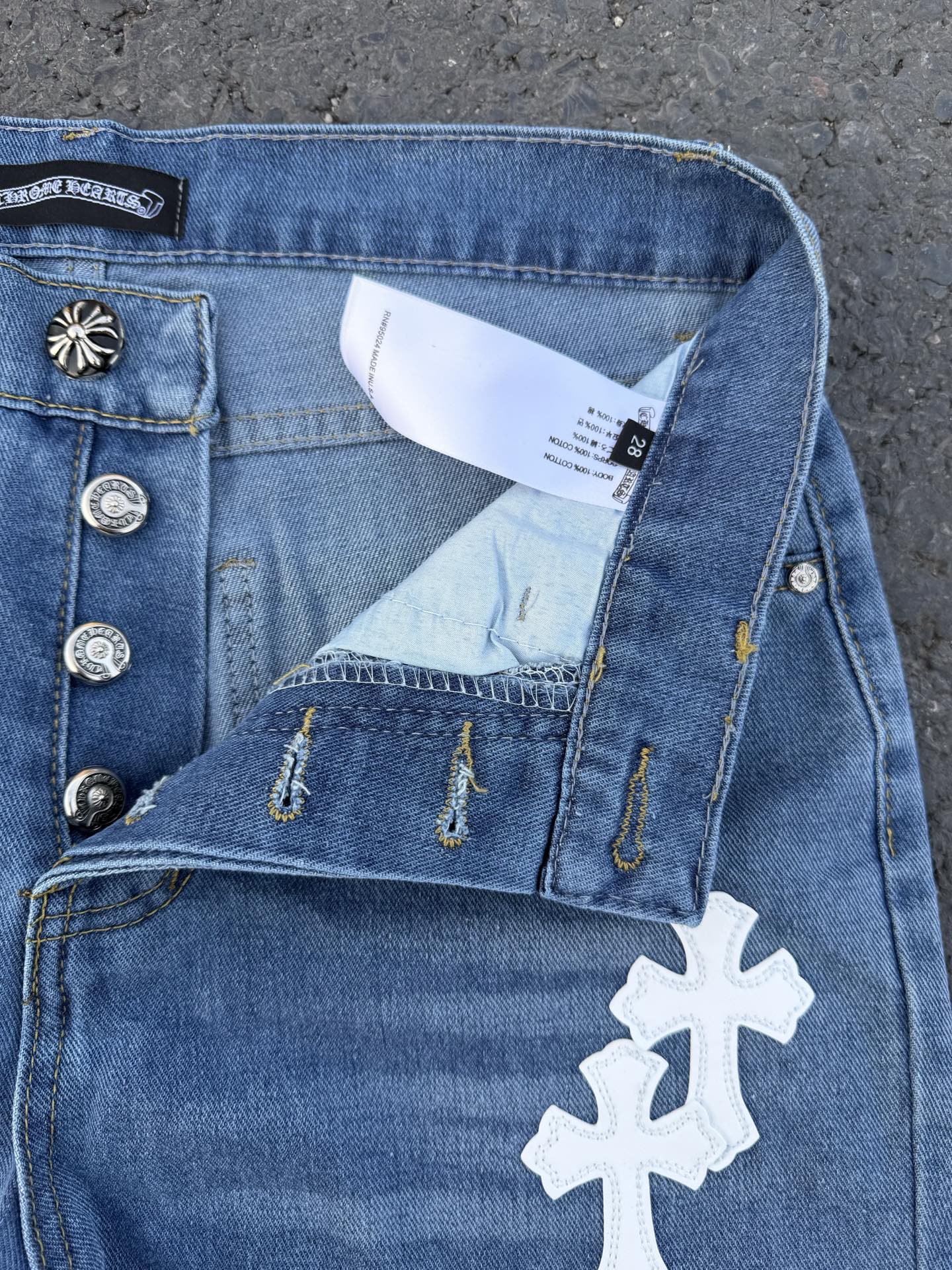 Light blue denim jeans with button fly, distress stitching, white cross patch.