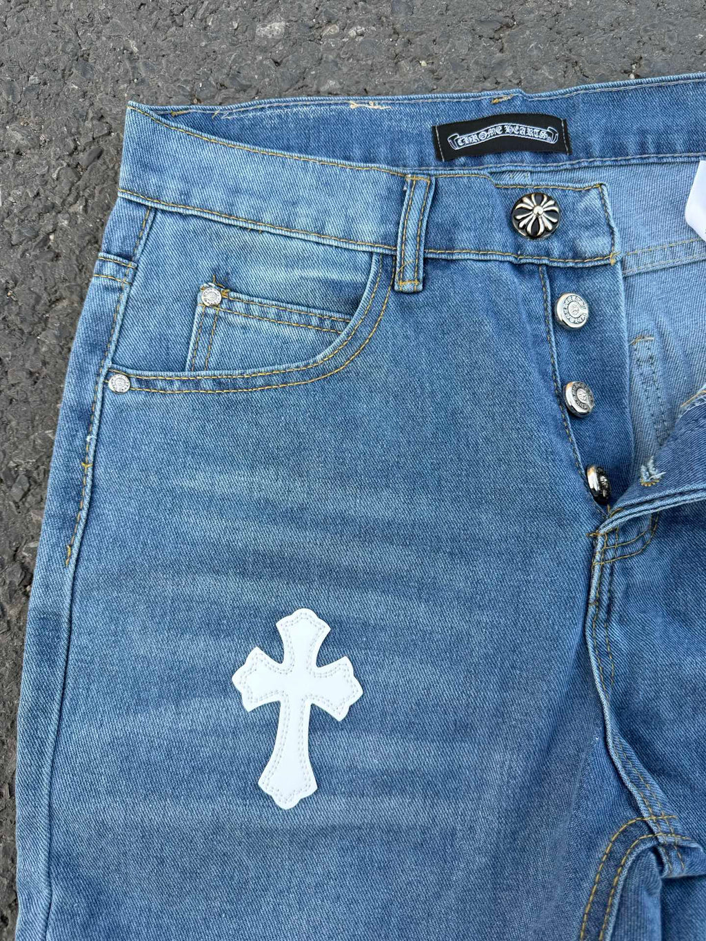 Light blue denim jeans with front pocket, white cross patch, button details.