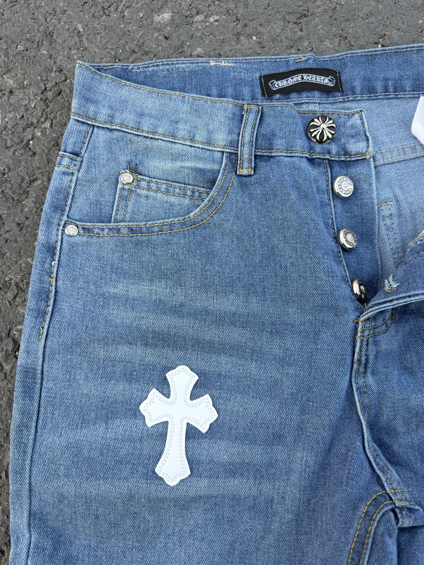 Light blue denim jeans with front pocket, white cross patch, button details.