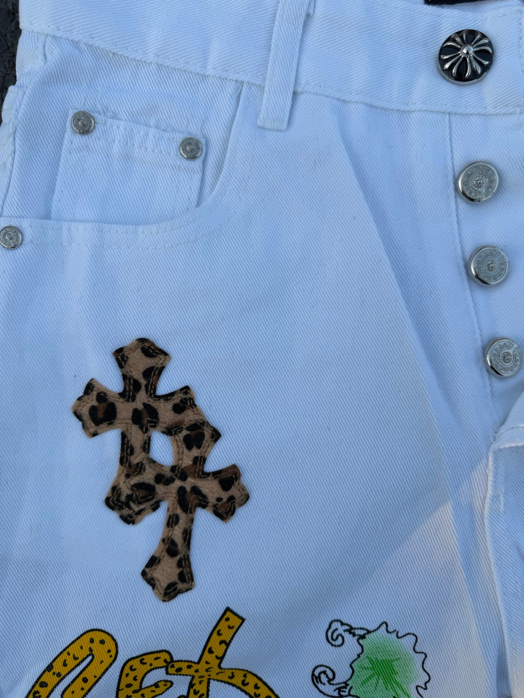 Light blue denim pants detail: metal buttons, button fly, and dark cross patch
