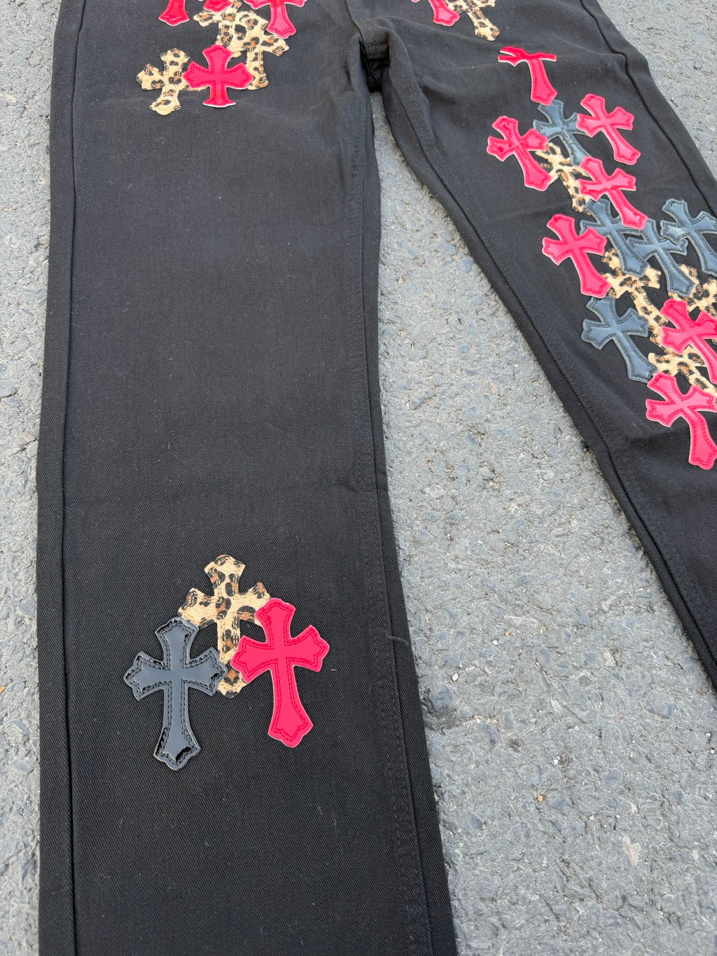 Black denim pants with scattered cross patches and multicolor script on the side.