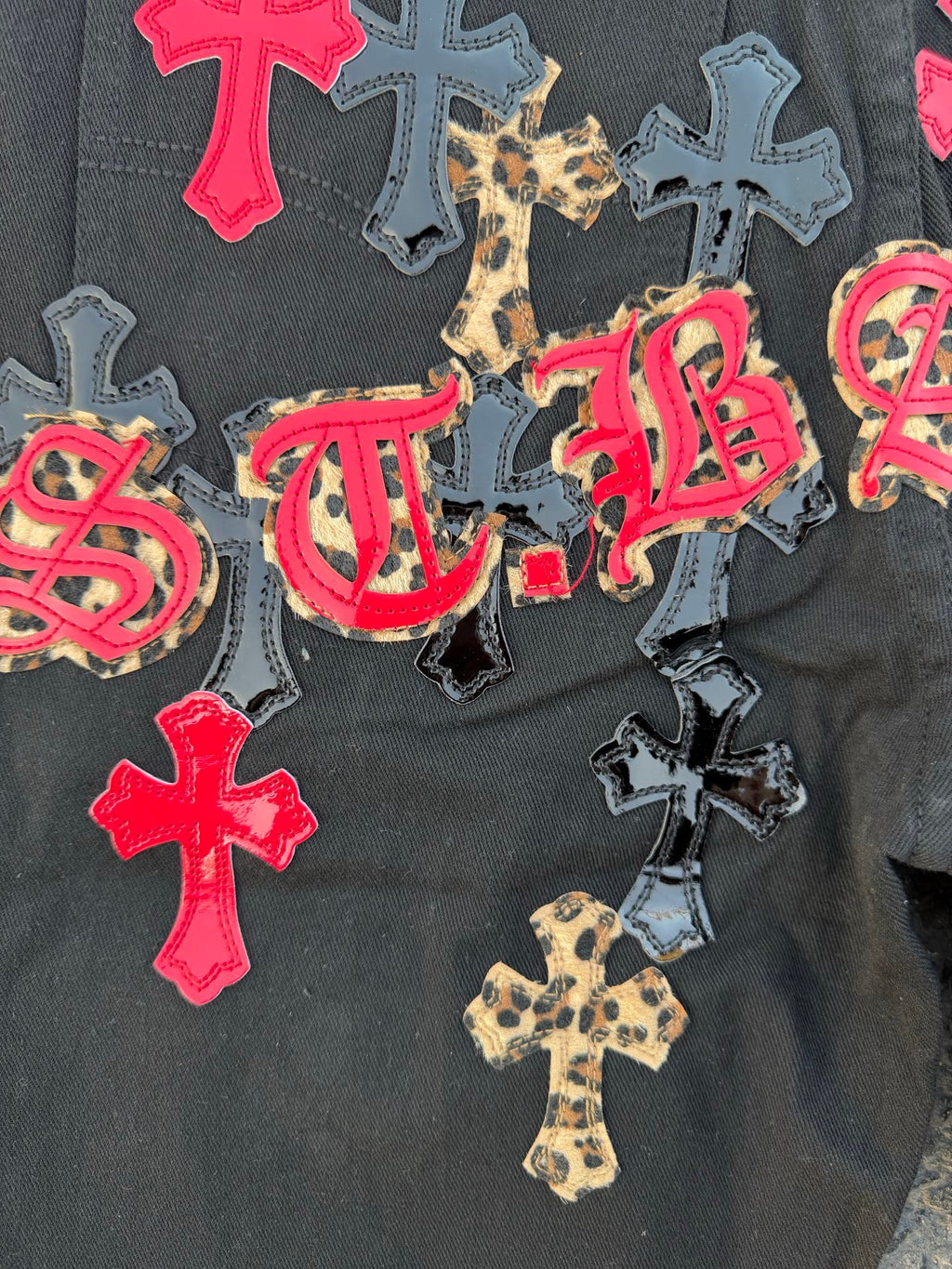 Black denim pants with colorful cross patches and script embroidery on the leg.