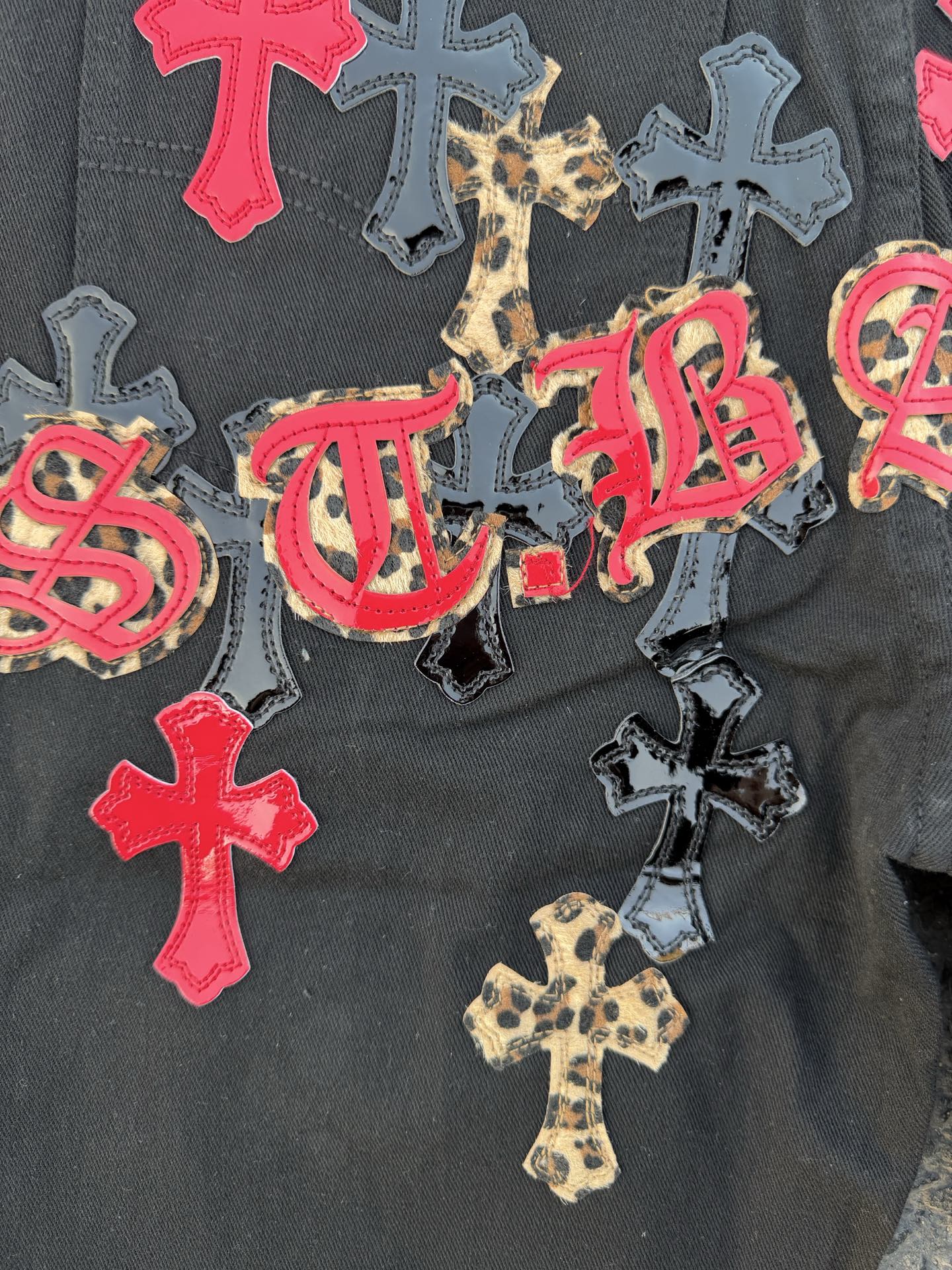Black denim pants with colorful cross patches and script embroidery on the leg.