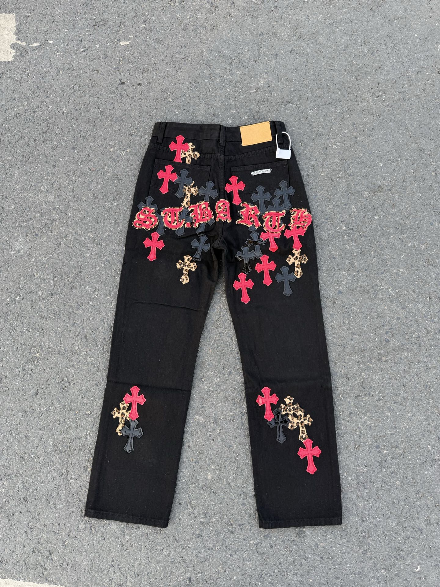 Black denim pants with colorful cross patches and a white tag on the waistband.
