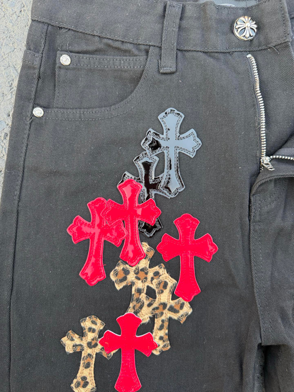 Dark gray denim pants with a zip fly and cross applique on the front pocket area.