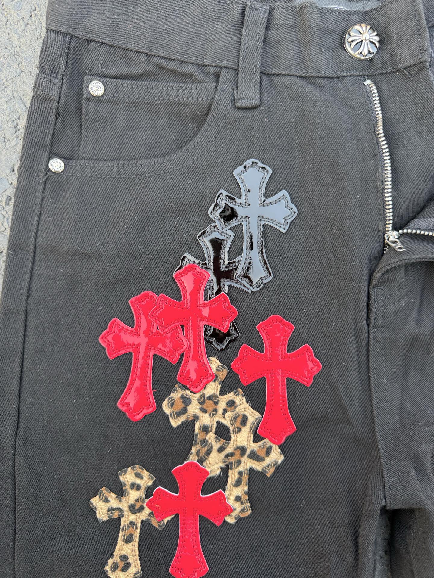 Dark gray denim pants with a zip fly and cross applique on the front pocket area.