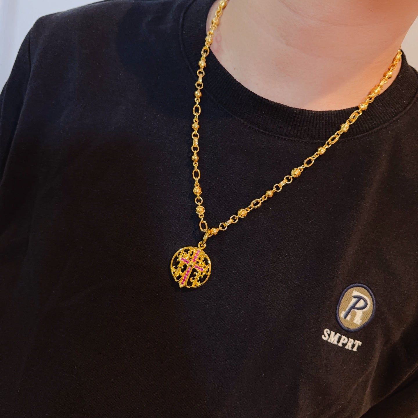 Gold-tone chain necklace with round red enamel crest pendant worn on dark T-shirt.