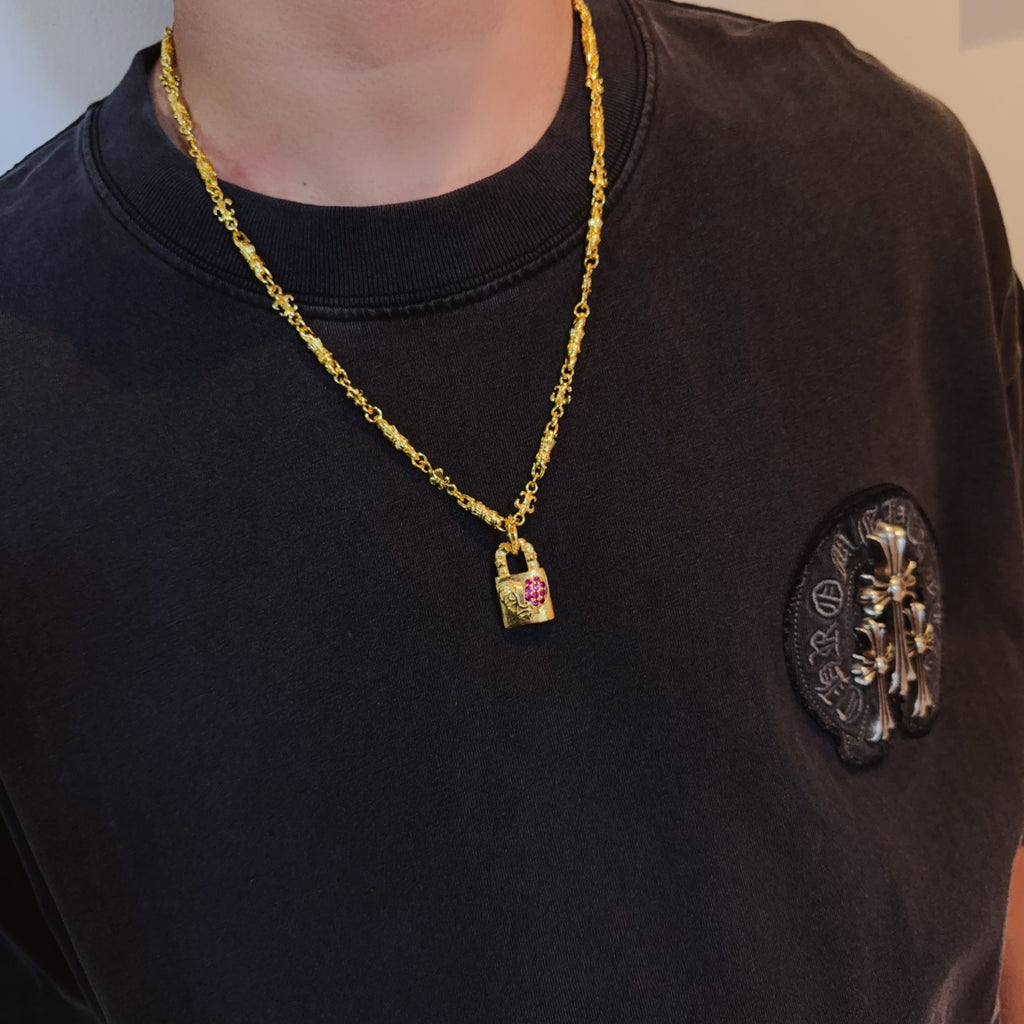 Gold-tone pendant necklace worn on dark shirt; small rectangular pink gemstone charm