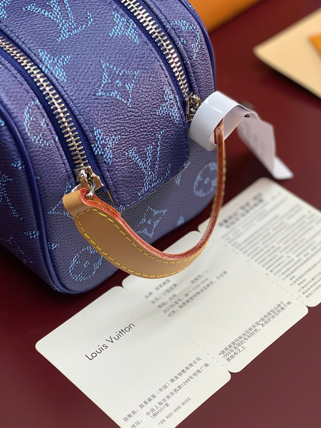 Blue monogram bag corner with gold-tone zipper, tan strap, and hanging white tag.