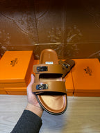 Hermes tan slide sandals held in hand, two strap buckles visible