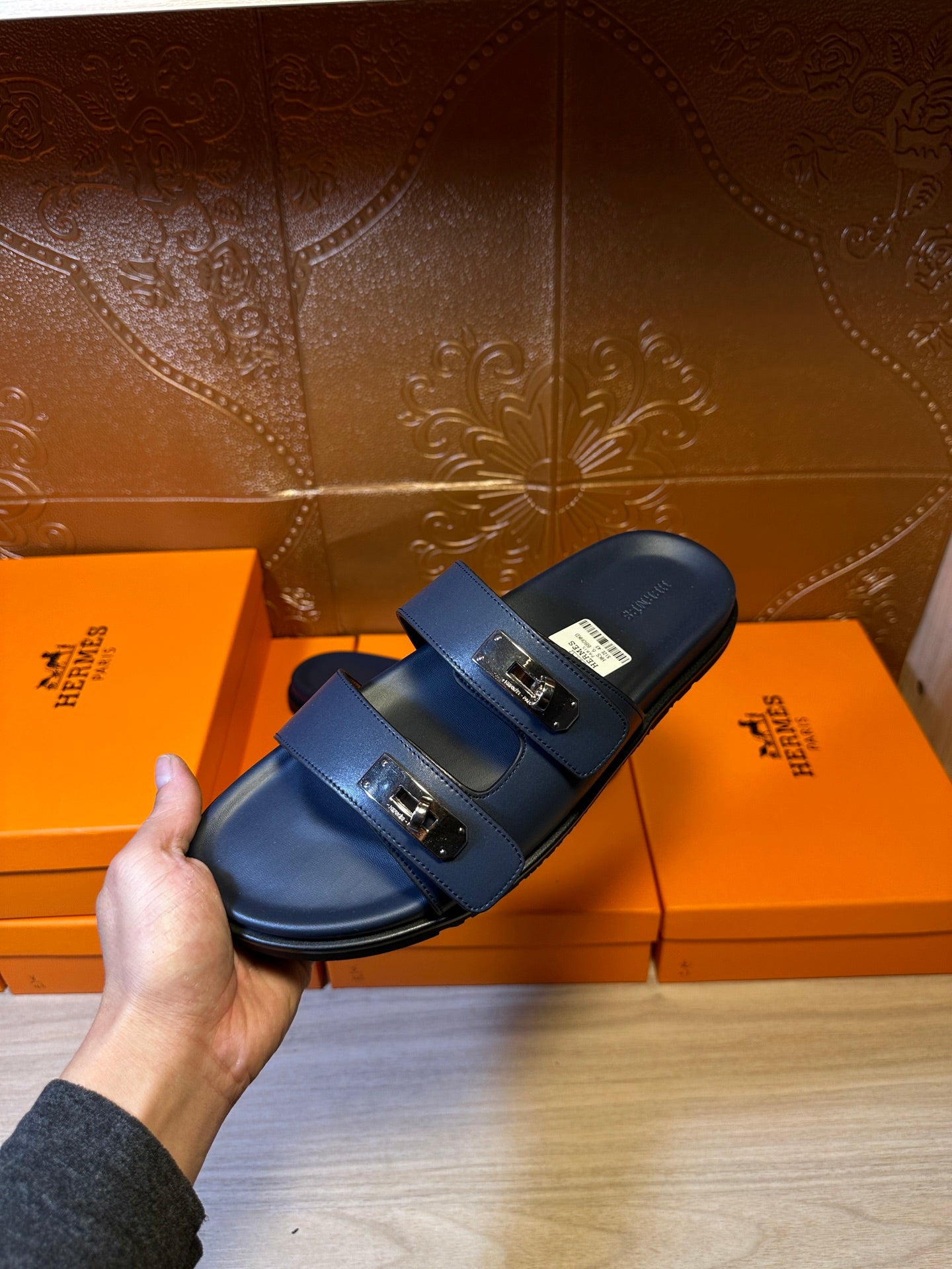 Hermes blue slide sandals with two front buckles, held in hand above boxes