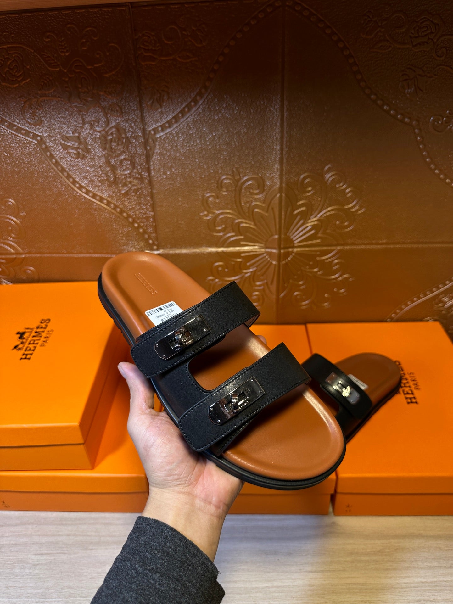 Hand holds a black two-strap slide sandal with silver buckles and tan insole