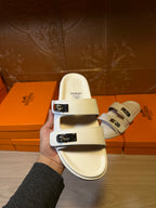 Hermes slide sandals, white upper; hand shows front view with metal buckle straps