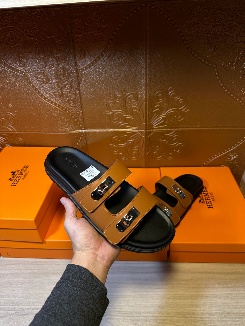 Tan and black slide sandals with silver buckle detail held over orange box