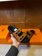 Tan and black slide sandals with silver buckle detail held over orange box