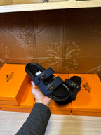 Hermes luxury sandals held in hand, navy straps with silver buckles on black footbeds.