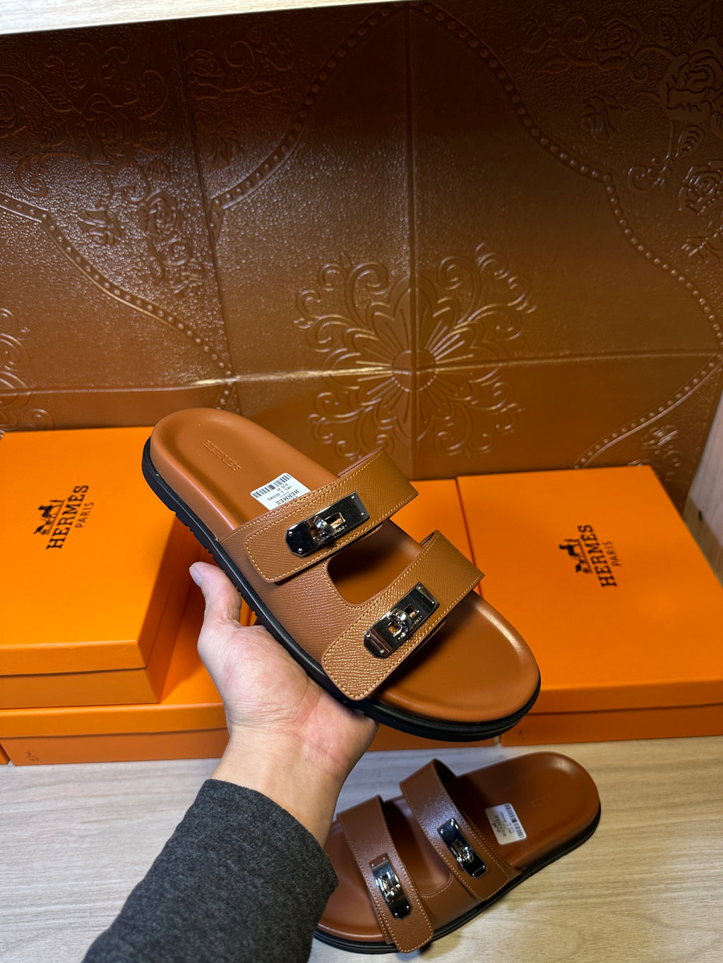 Tan leather slide sandals with silver buckles and open toe, hand holding one pair