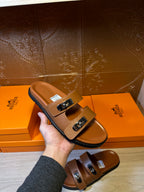 Tan leather slide sandals with silver buckles and open toe, hand holding one pair