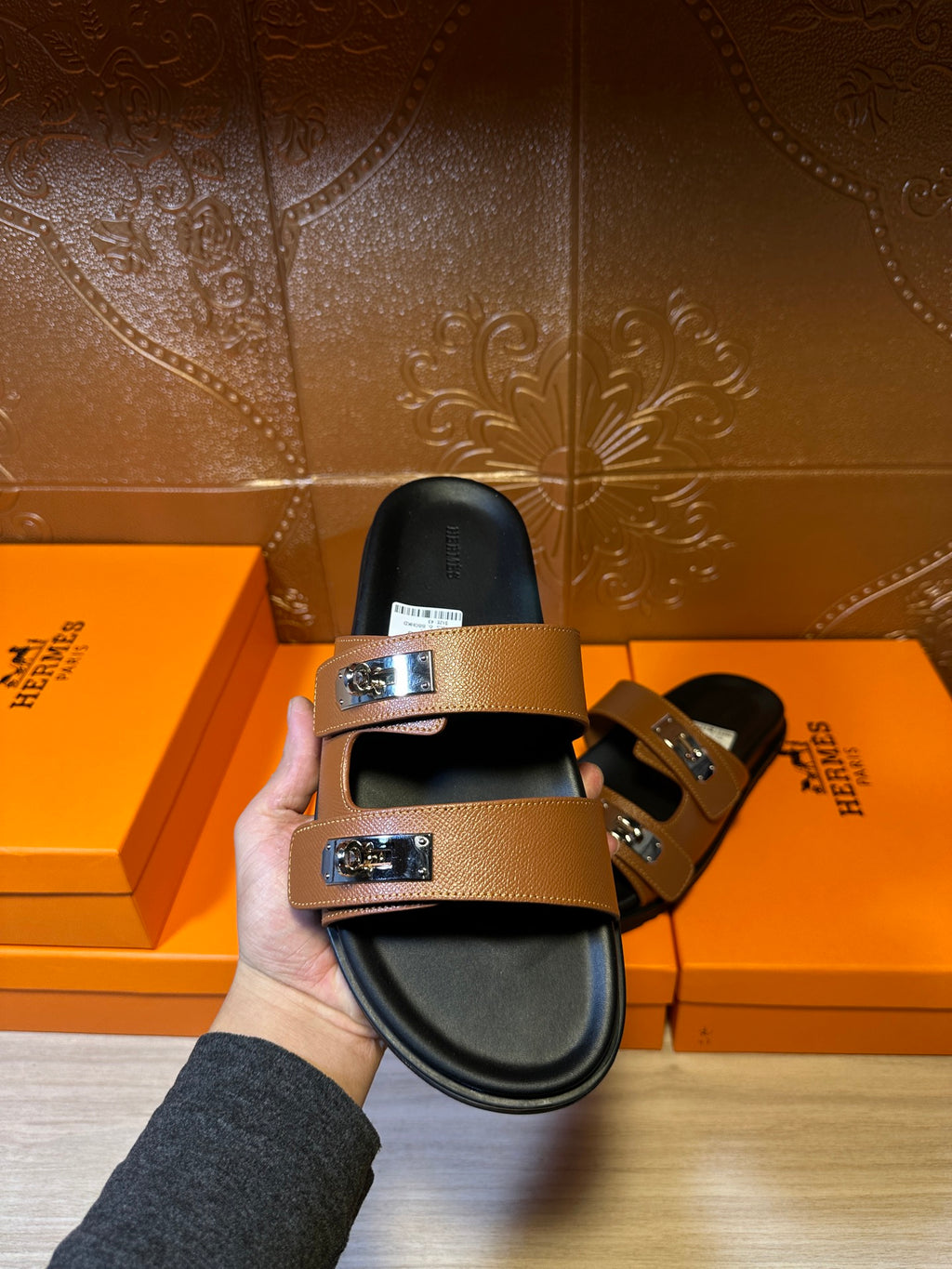 Tan leather sandals held up, featuring two metal buckle straps over a black footbed