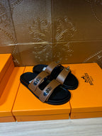 Tan slide sandals with two wide straps, black footbed, and rectangular metal buckle plates