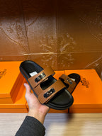 Hand holding tan sandals showing black footbed and two silver buckle straps