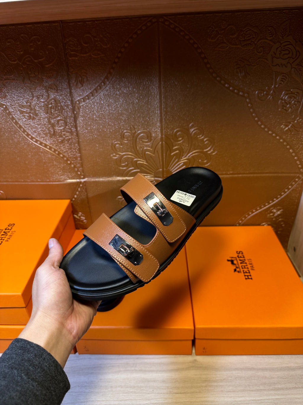 Tan sandals held in hand, showing double straps and silver rectangular buckle hardware