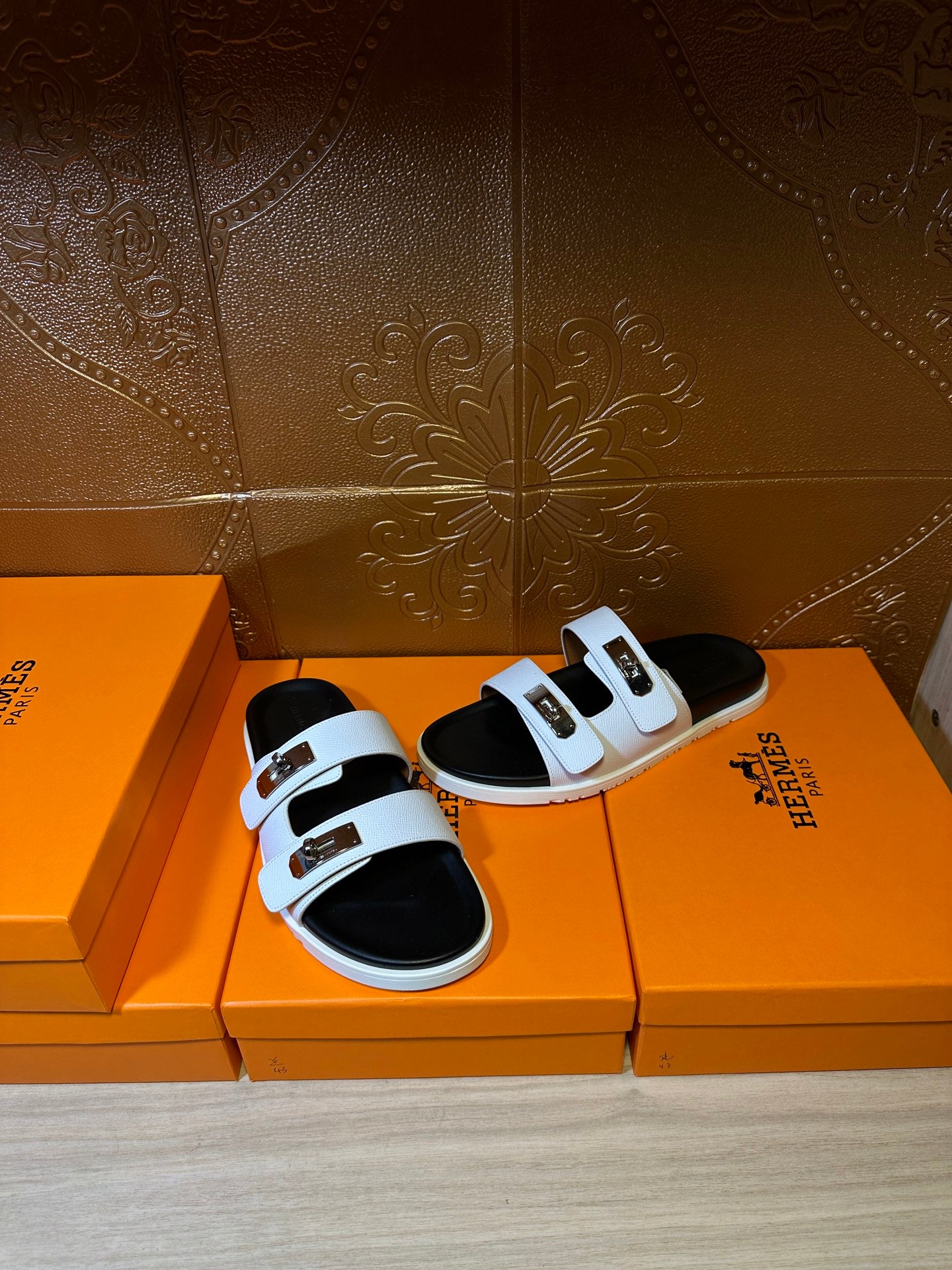Hermes slide sandals set on orange boxes, white straps with gold buckles and black footbeds