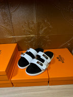 Hermes slide sandals angled view, white straps over black footbeds with gold buckles