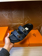 Hand holding Hermes blue slide sandal, front strap with two silver buckles
