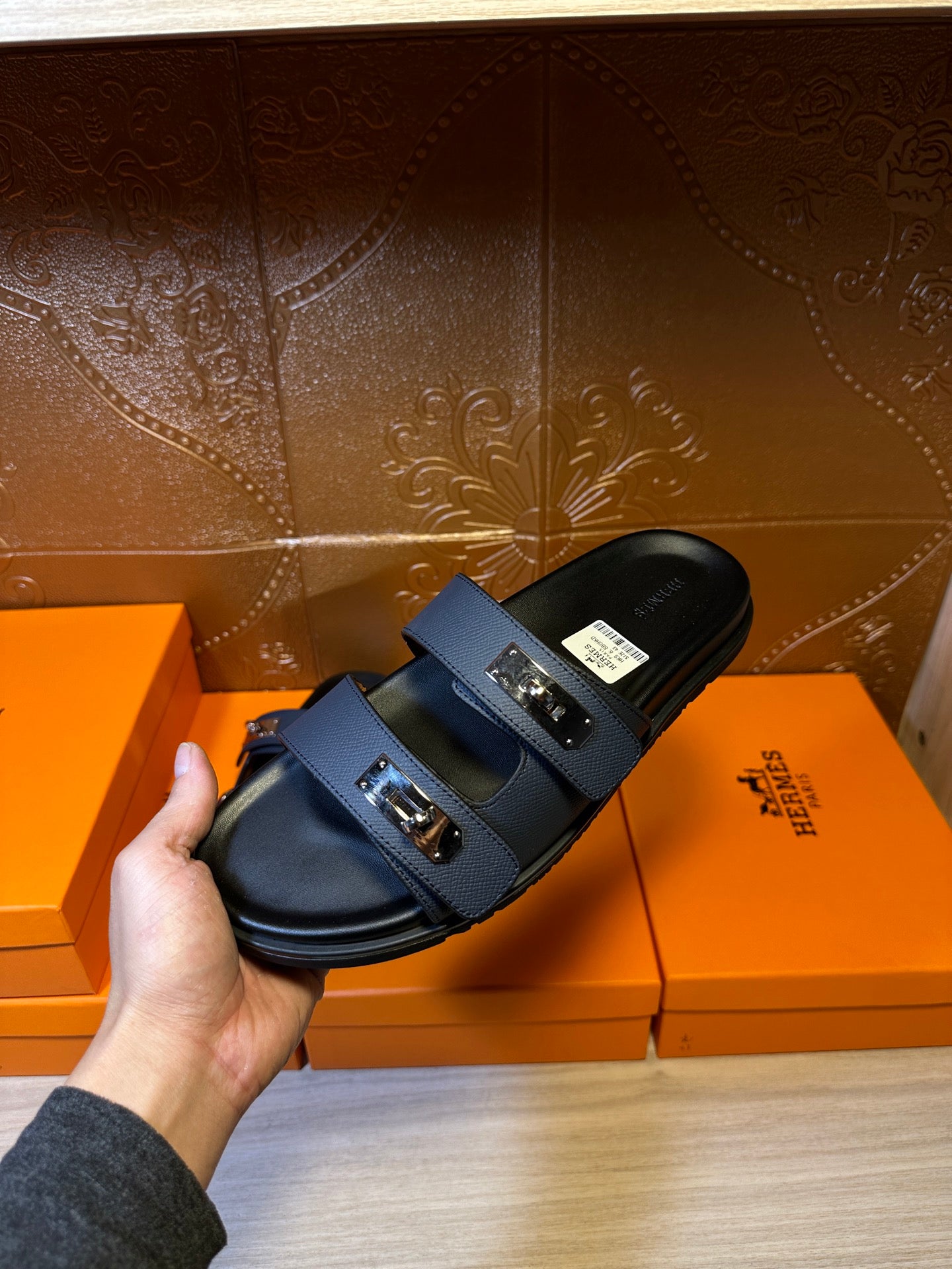 Hand holding Hermes blue slide sandal, front strap with two silver buckles