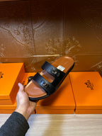 Dark slide sandals held above box; two straps with buckle hardware