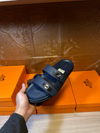 Blue leather slide sandals with two metal buckle straps held above display boxes