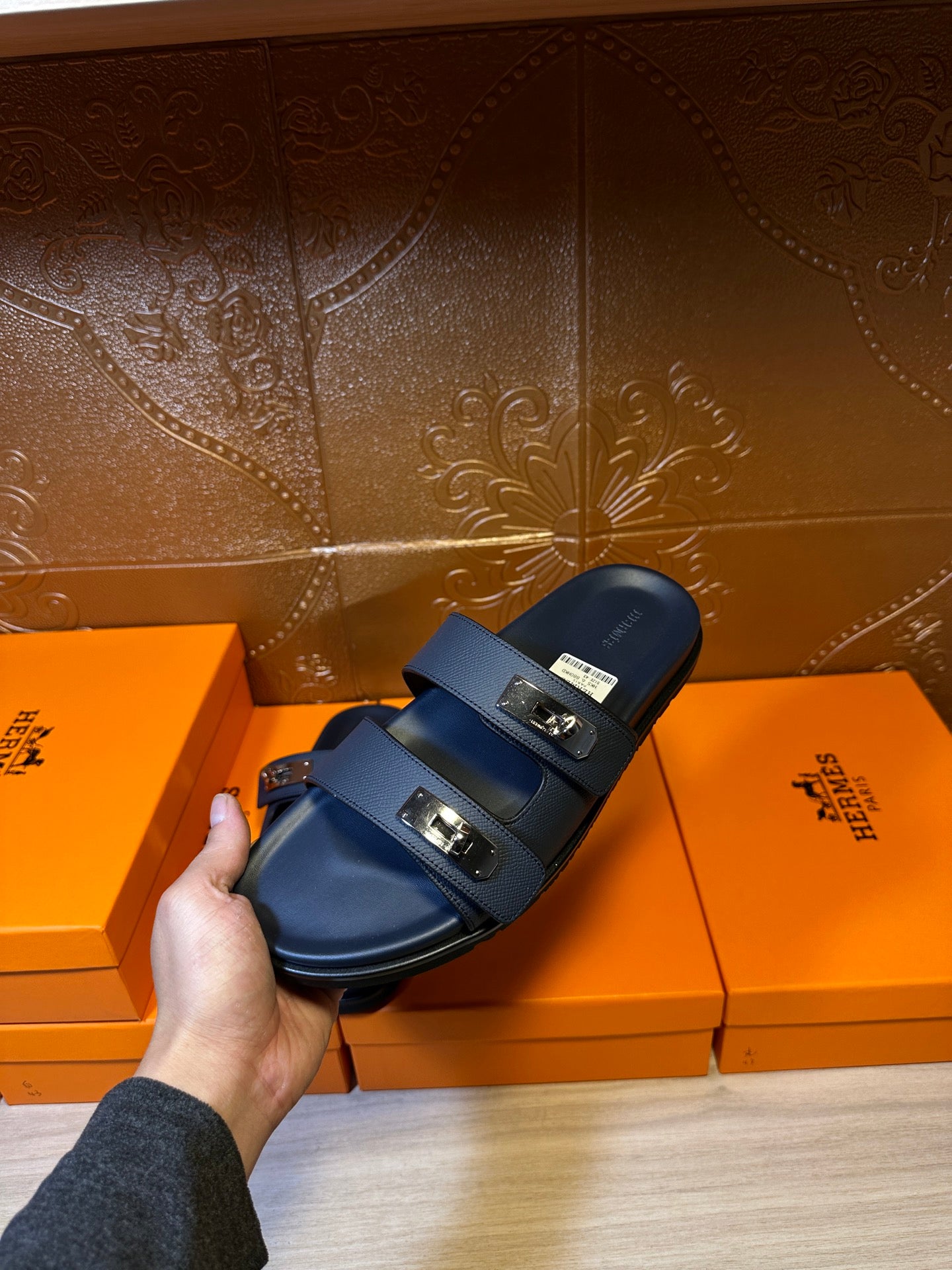 Blue leather slide sandals with two metal buckle straps held above display boxes
