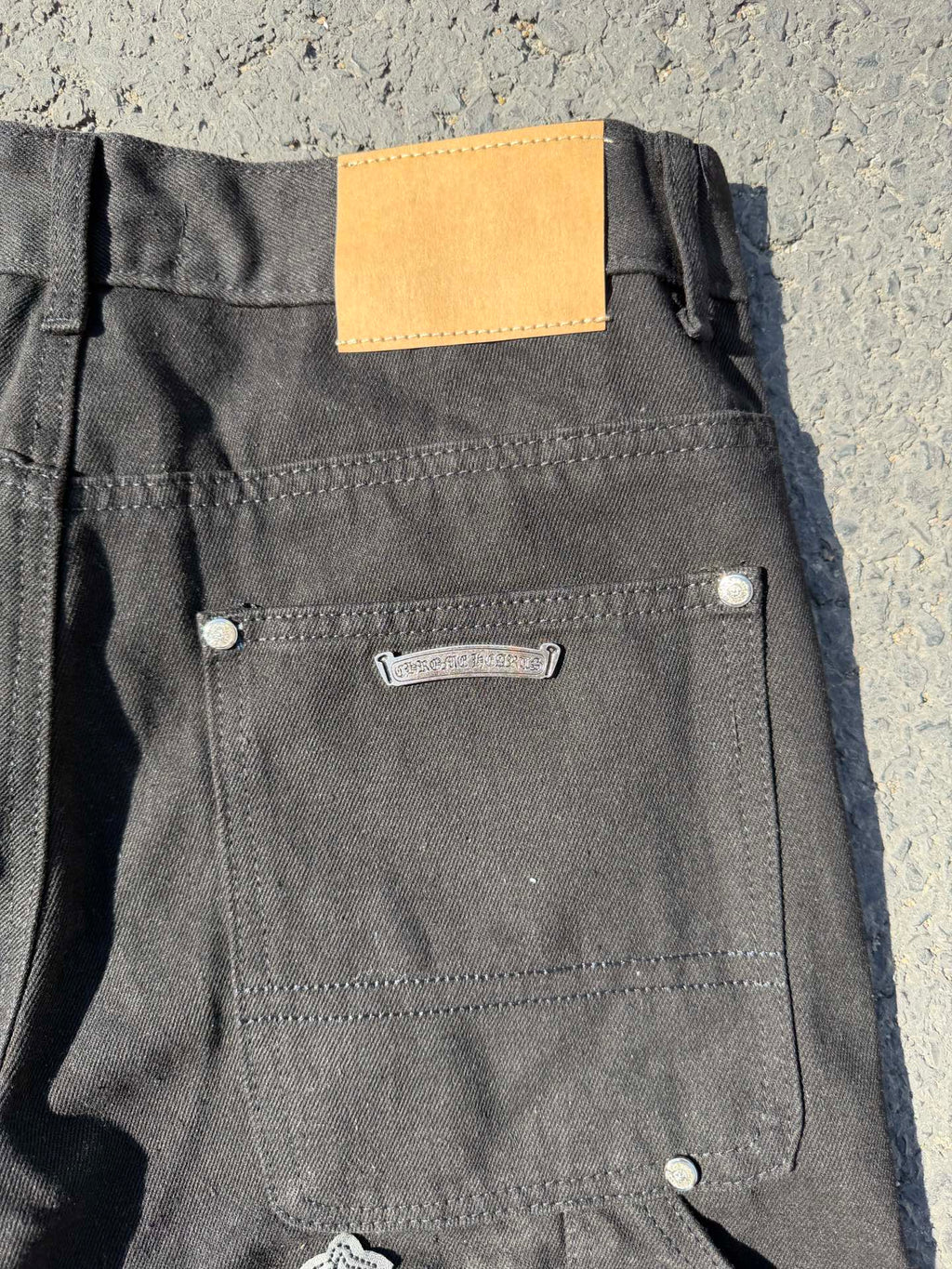 Black cargo pants with back pocket, metal button, and small cross patch near hem