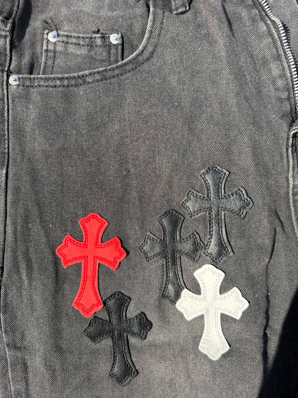 Close-up of dark gray denim pants with red, white, and black cross embroidery