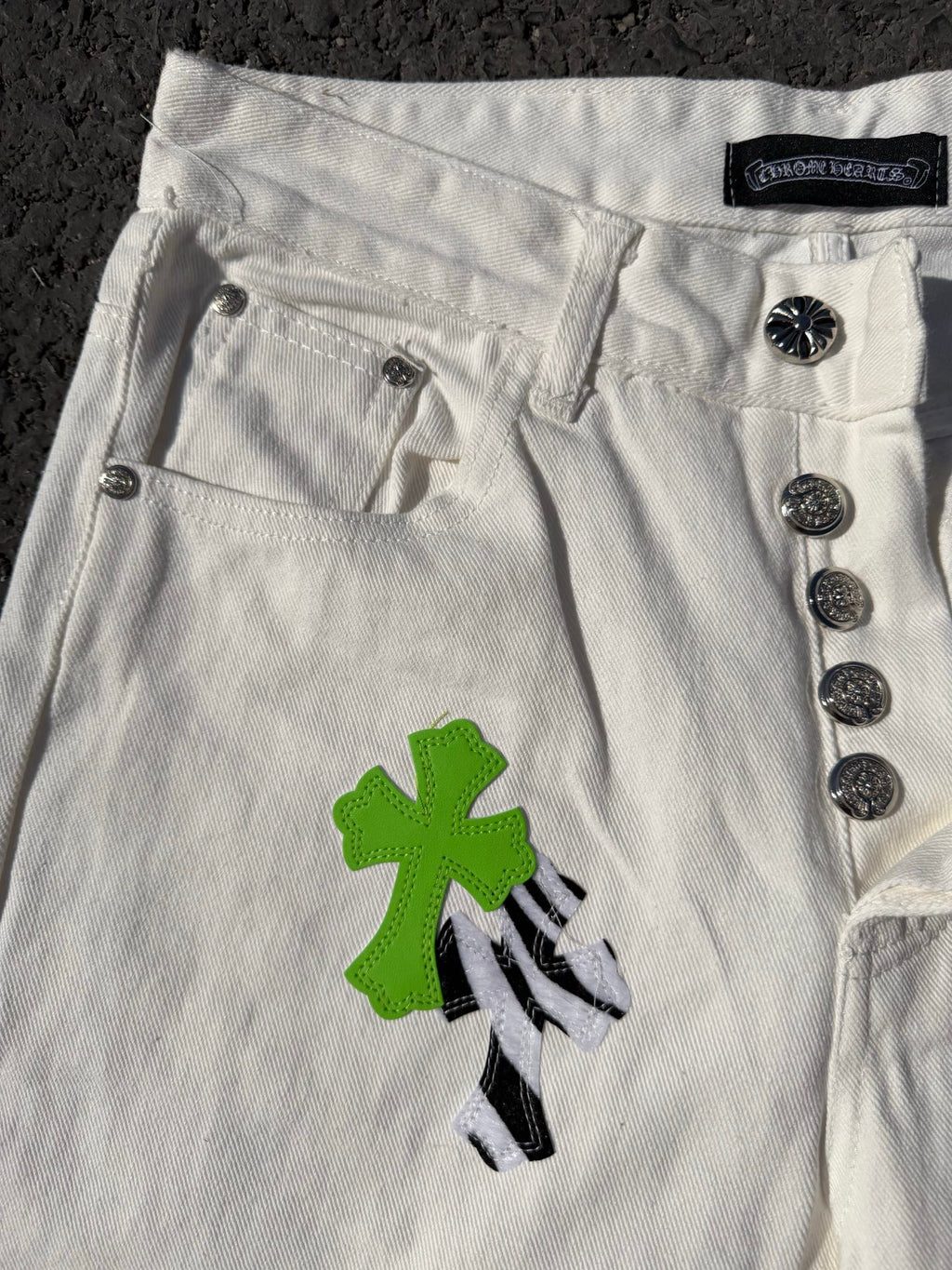 White denim pants close-up of buttoned waistband and green cross applique with black detail