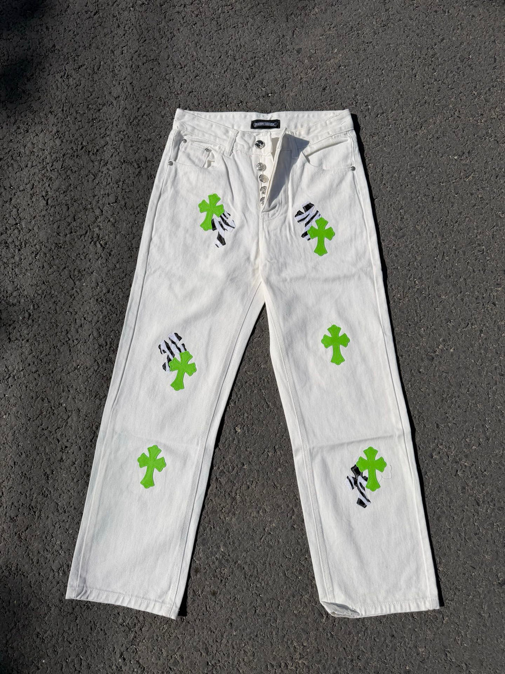 White denim pants with button fly, green cross patches, and black graphic details